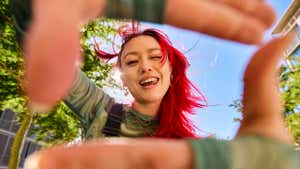 Young adult with red hair smiling and looking through a hand frame gesture outdoors in an urban environment. Low angle shot.