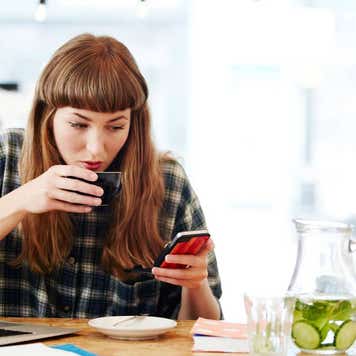 Person working at table with computer, drinking coffee and looking at smartphone